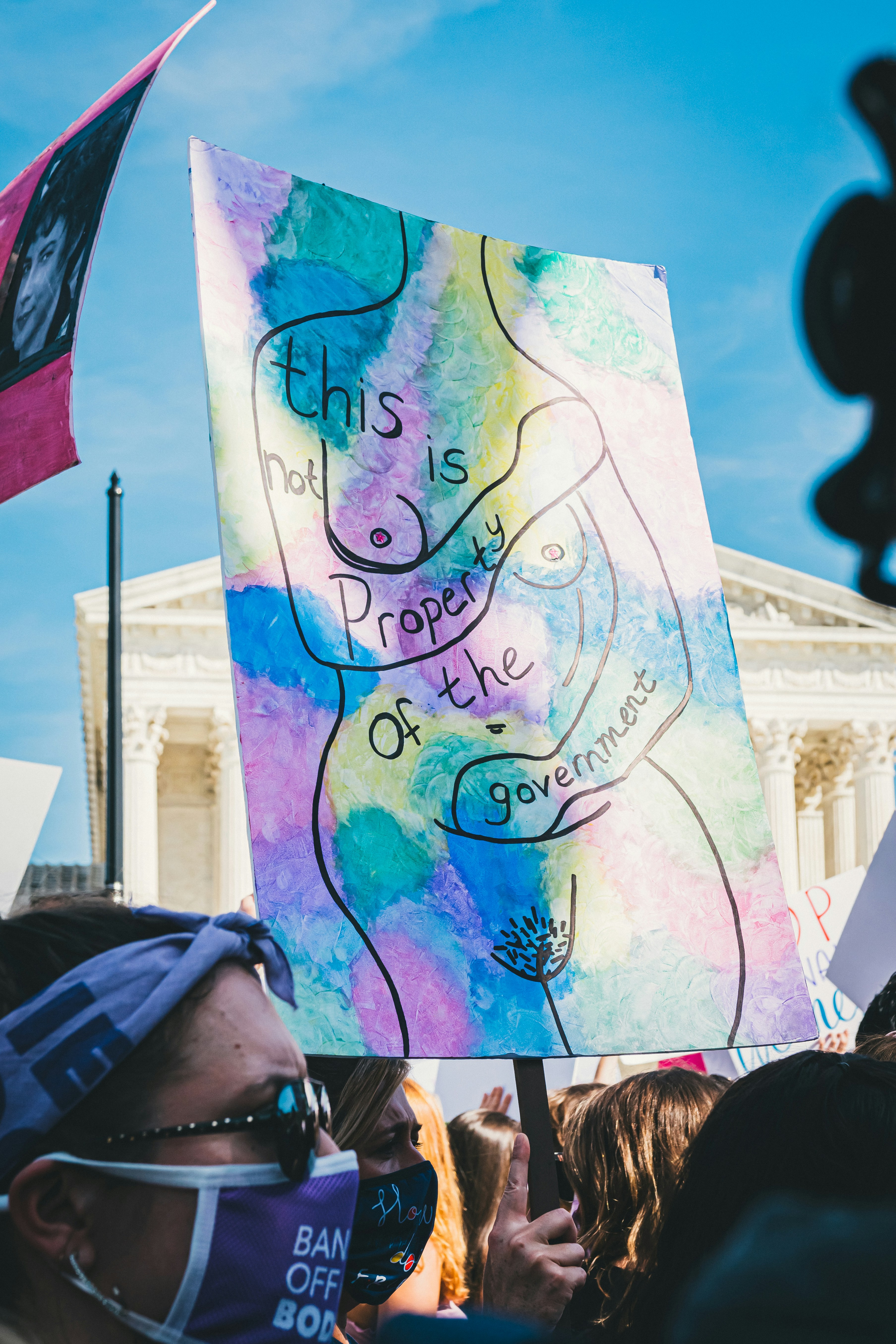 Das Bild zeigt eine Demonstration, bei der eine Person ein Schild hält, auf das ein nackter Torso gezeichnet ist, mit der Aufschrift "This is not property of the government"