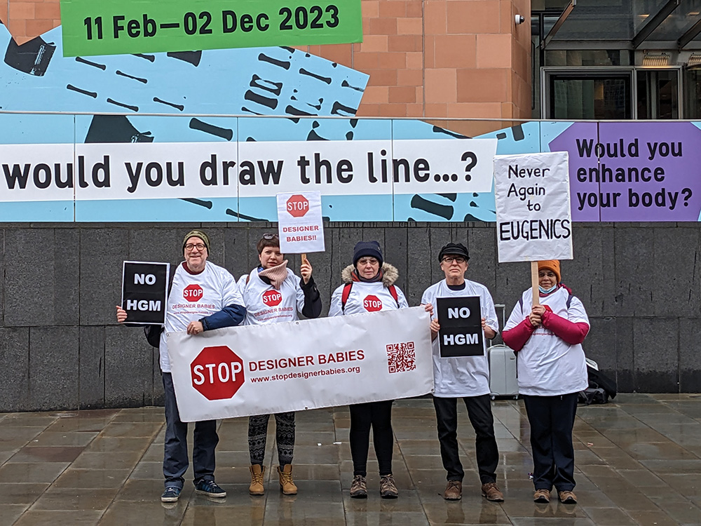 Fünf Menschen stehen vor einem Gebäude. Sie halten Schilder und ein Banner mit der Aufschrift "Stop Designer Babies".