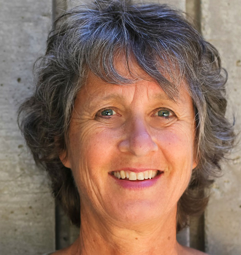 Porträt einer älteren Frau mit kurzen, welligen grauen Haaren. Sie lächelt leicht und blickt direkt in die Kamera. Ihr Gesicht ist warm ausgeleuchtet, im Hintergrund ist eine graue Betonwand zu sehen.