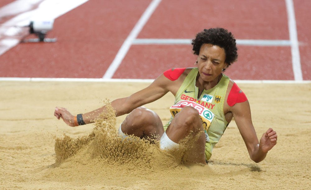 Eine Leichtathletin landet nach einem Weitsprung im Sand. Sie sitzt leicht rückwärtsgelehnt im Sandbett, der helle Sand spritzt um sie herum. Sie trägt einen sportlichen Einteiler in Gelb- und Rottönen mit der Aufschrift „Germany“. Im Hintergrund ist die rote Laufbahn der Sprunganlage zu sehen.