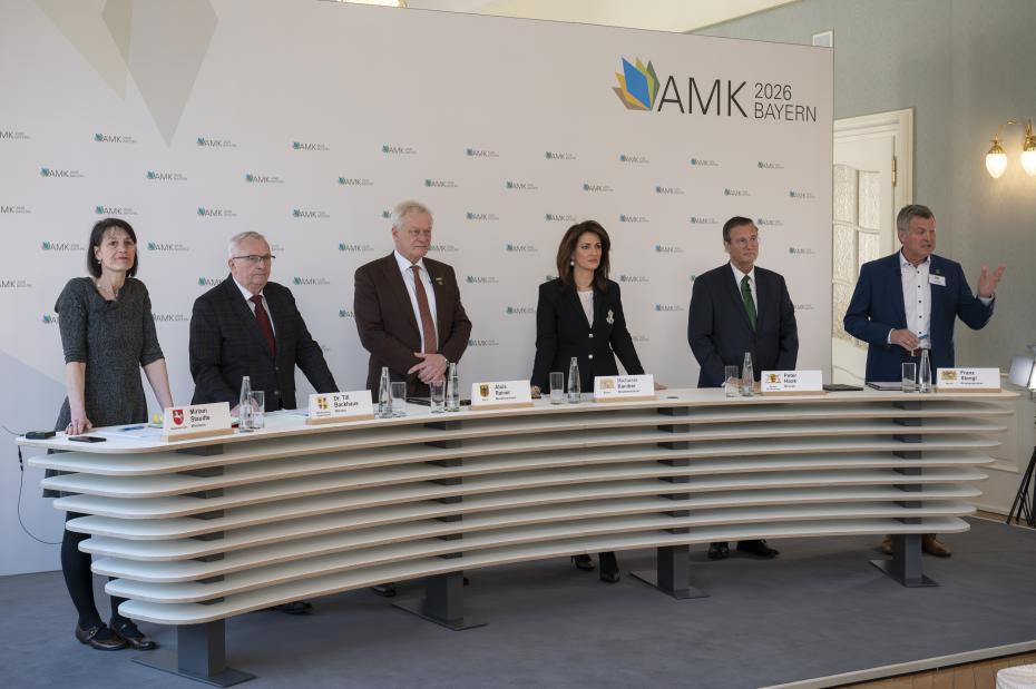 Fünf Personen stehen hinter einem langen, geschwungenen Pult mit Namensschildern und Wassergläsern, im Hintergrund eine Wand mit dem Schriftzug 'AMK 2026 BAYERN'