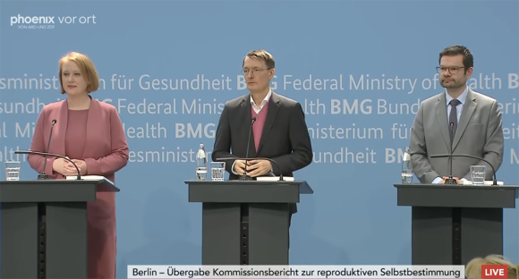 Screenshot aus dem Video zur Live-Übertragung der Übergabe des Kommissionsberichts. Zu sehen sind die Minister*innen Paus, Lauterbach und Buschmann. Sie stehen jeweils hinter Redner*innenpulten vor einem blauen Hintergrund.